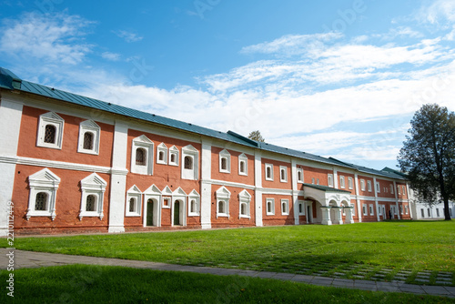 A historic building made of red bricks stands in a grassy area. The sky is clear with some clouds.