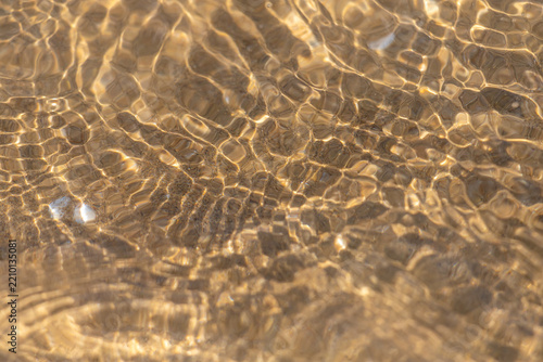 Golden sunlight ripples over shallow sandy water creating abstract shimmering...