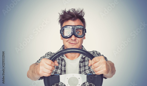 Smiling man wearing motorcycle goggles grips a car steering wheel from the front, symbolizing driving, adventure, speed, confidence, road travel and playful automotive enthusiasm and humor!