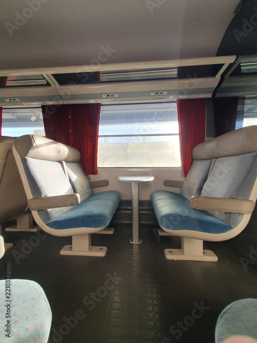 Empty Train Interior With Paired Seats and Window Table, Sunlit Commuter Carriage With Red Curtains