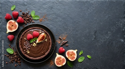 Delicious homemade chocolate tart decorated with fresh figs and ripe strawberries on a dark rustic stone background