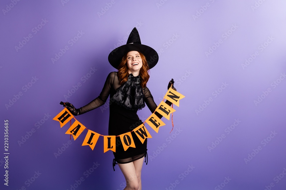 Obraz premium Young woman in witch costume holding Halloween banner against purple background, smiling and celebrating seasonal costume party