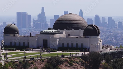 Aerial panning footage captures Griffith Observatory on Mount Hollywood in Griffith Park, with tourists walking along the terrace while the downtown Los Angeles skyline rises in the distance.