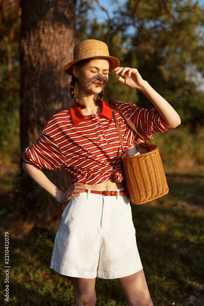 Obraz premium Young woman in a straw hat and striped shirt with a wicker bag poses outdoors during golden hour sunlight, enjoying a summer lifestyle while standing relaxed in a sunlit forest glade.