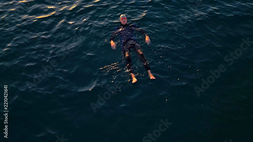 Obraz na plátně An athlete in a black wetsuit and red goggles floats in the deep blue ocean, practicing open-water swimming with a focused expression, highlighting determination and challenge in sport training