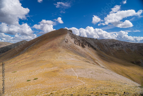 View of San Luis Peak with a hiking trail winding up the mountain slope under a blue sky with white clouds in Creede, Colorado, United States.
