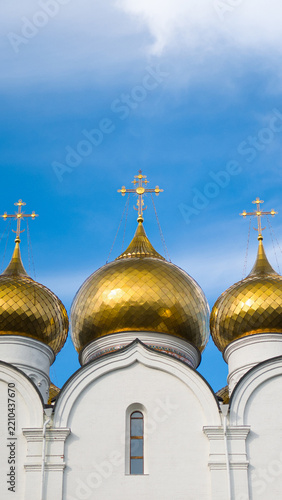 A Dormition Cathedral stands with golden domes under a clear blue sky. The entrance features a colorful mural and steps leading up to the main door.