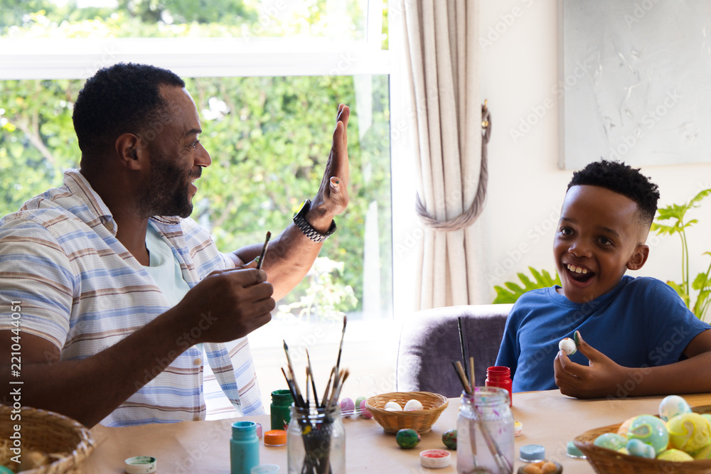 Obraz premium African American father and son painting eggs at wooden table by window, holding paintbrushes