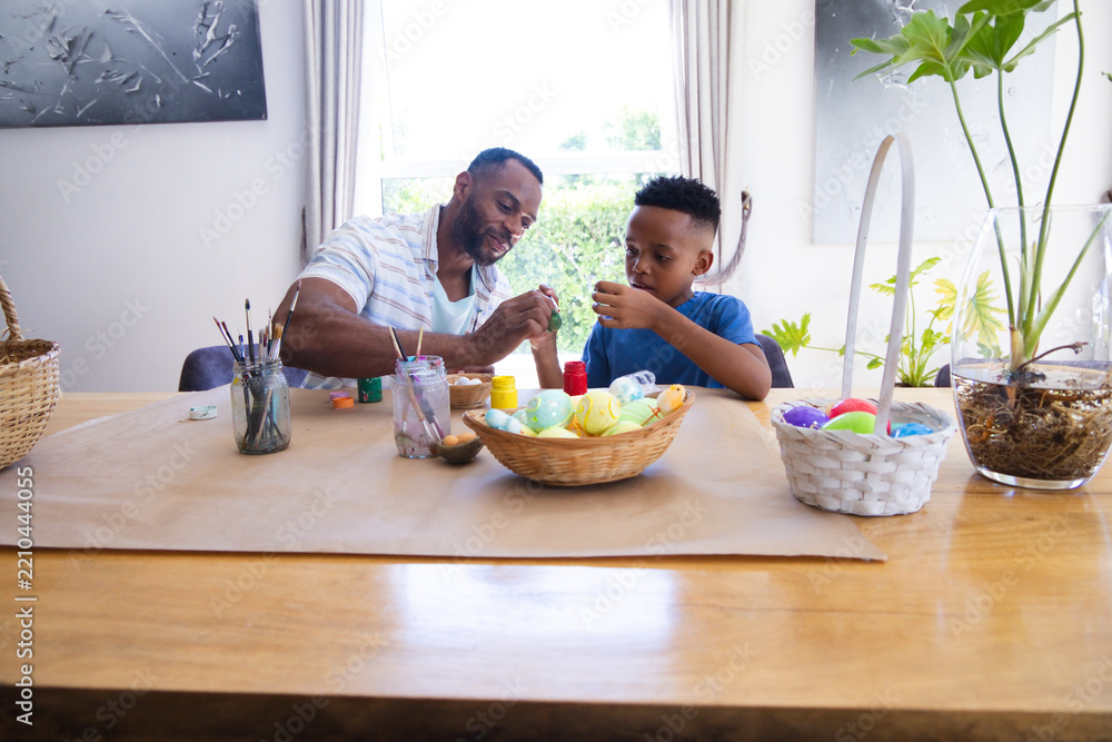 Obraz premium African American father and child painting colorful Easter eggs at dining table with paint jars