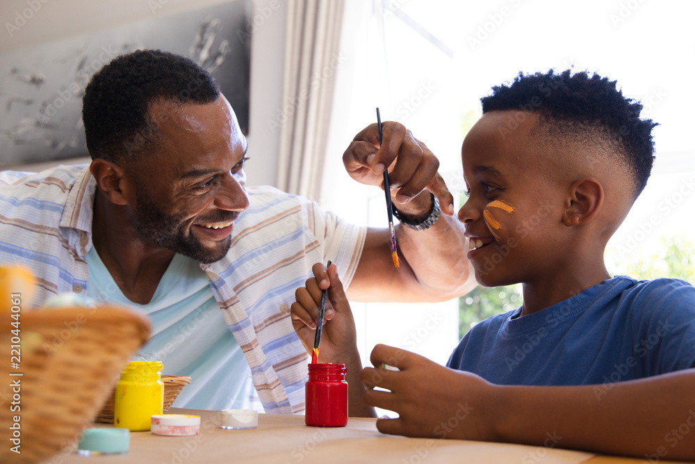 Obraz premium African American father in striped shirt and son painting at home table with paintbrushes and jars