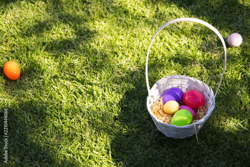 White woven basket is sitting right on sunlit grassy lawn holding colorful plastic eggs in straw
