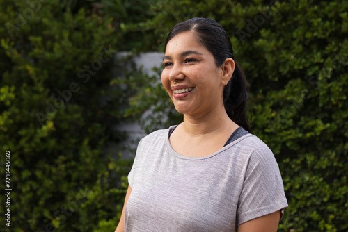 Woman standing smiling looking left in backyard with light gray shirt, dark straps, ponytail, hedge
