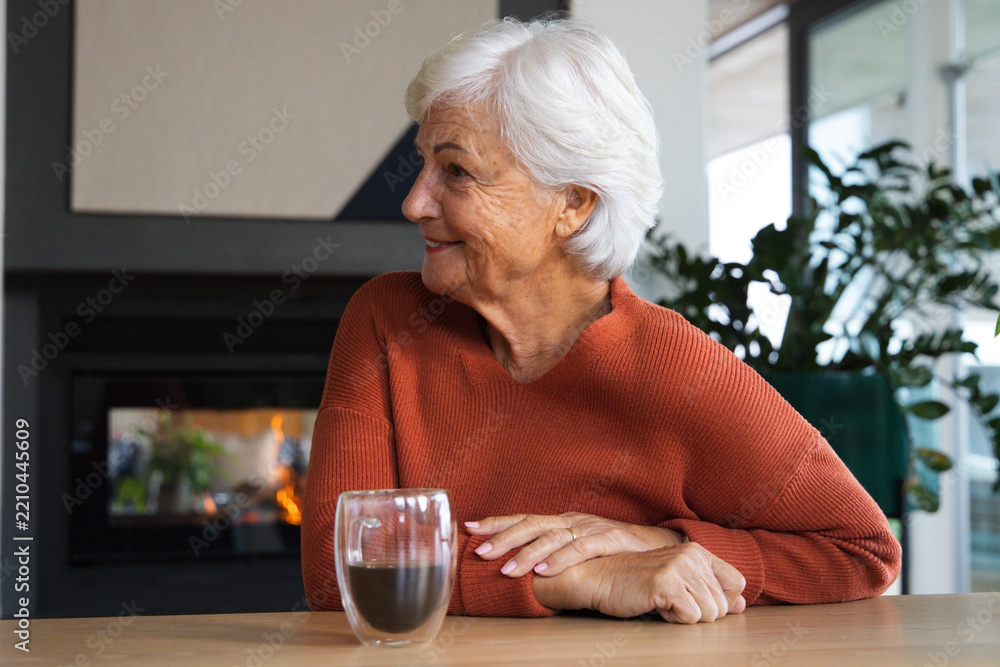 Fototapeta premium Senior woman sitting at table in lounge wearing rust sweater, holding glass mug, fireplace glowing