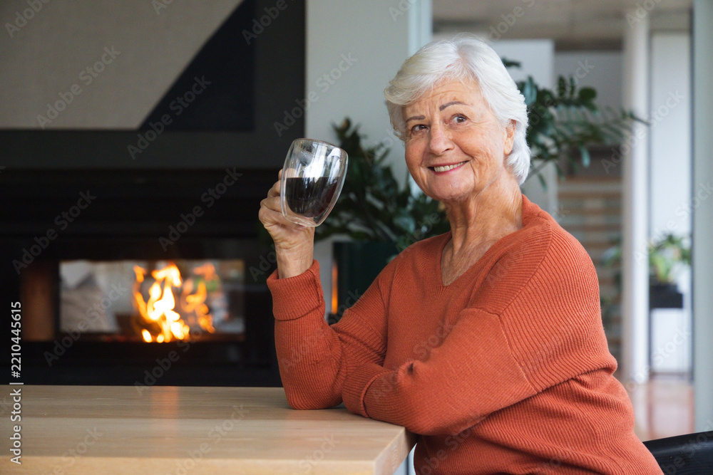 Obraz premium Senior woman holding glass mug in living room by lit fireplace wearing rust sweater, copy space