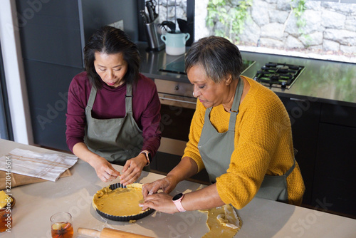 Asian, African American cooks shaping pie crust on kitchen island in olive aprons using rolling pin