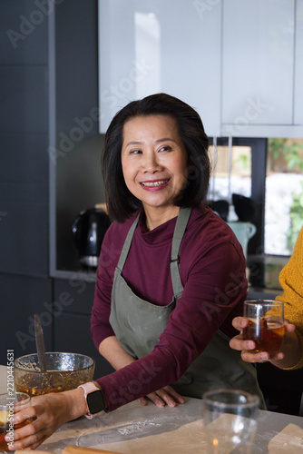 Asian friends watching mature woman preparing batter at kitchen island with mixing bowl and glass