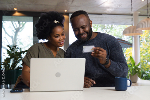 African American couple sitting at white table using laptop at home holding credit card and phone