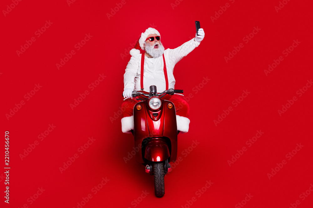 Obraz premium Santa Claus on a red scooter taking a selfie while wearing sunglasses and a festive costume against a red background