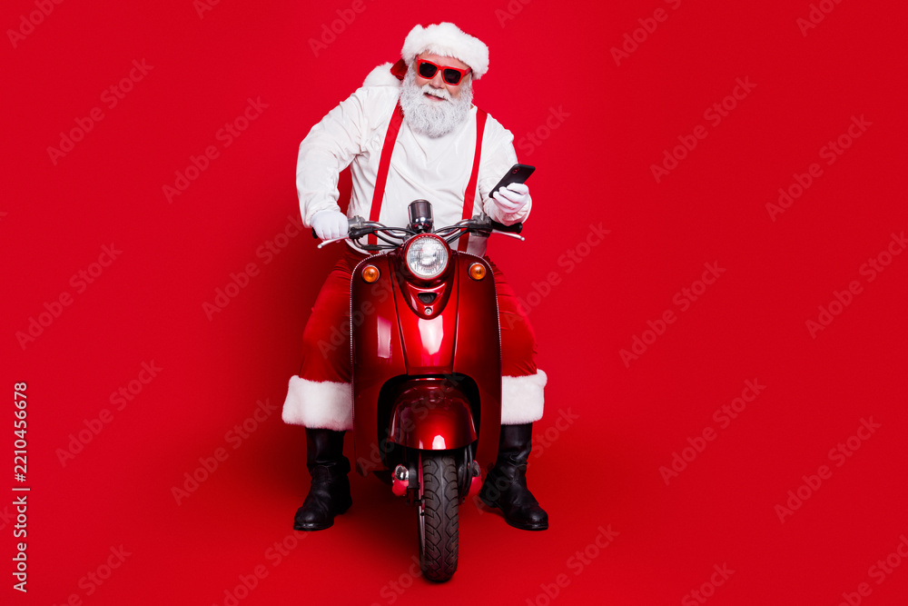 Obraz premium Santa Claus on a red scooter wearing sunglasses and holding a smartphone for modern Christmas delivery and holiday fun