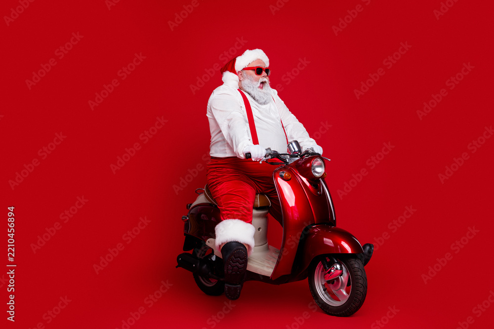 Obraz premium Santa Claus in red suit riding a shiny red scooter for Christmas celebration and holiday fun, playful festive portrait with white beard and sunglasses on vibrant red background