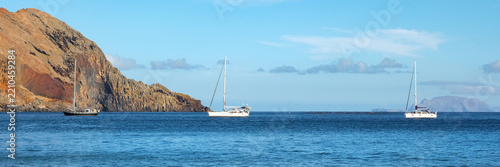 Three sailboats anchored in the calm blue ocean near rugged volcanic cliffs under a clear sky, creating a scenic coastal travel landscape