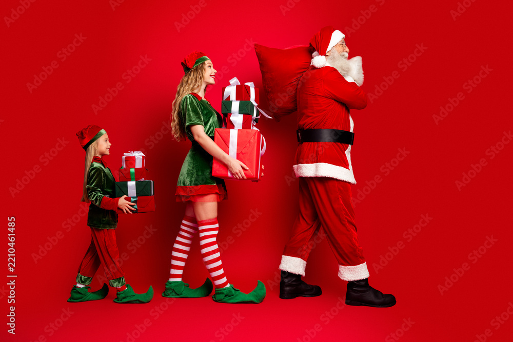 Obraz premium Santa Claus and two festive elves carrying wrapped Christmas presents in a cheerful holiday procession against a red background