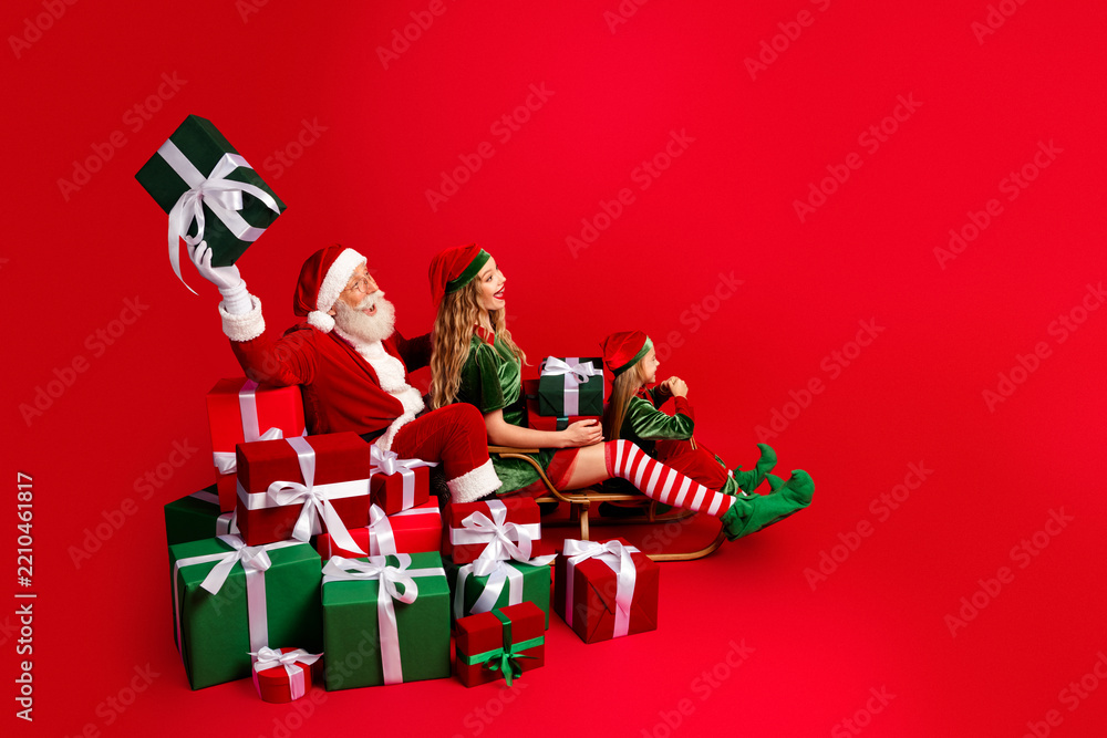 Obraz premium Santa Claus and two cheerful elves with wrapped Christmas gifts on a sled against a bright red background celebrating holiday joy