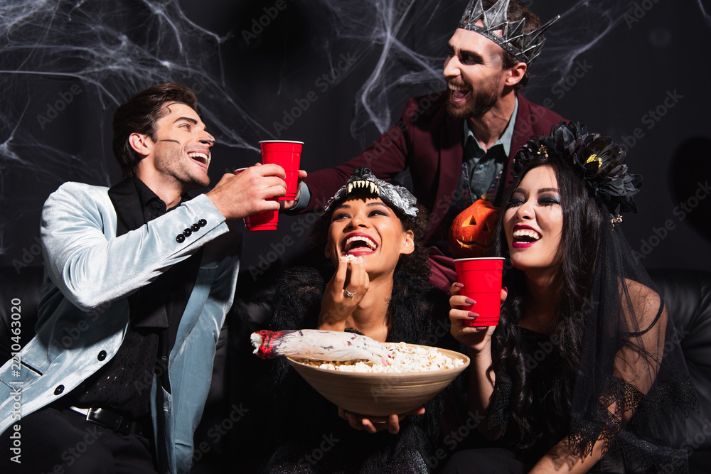 Obraz premium Multicultural friends joyfully celebrating Halloween in costumes with drinks and popcorn 