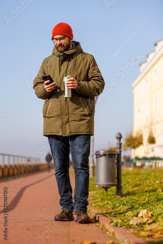 Man taking a walk using smart phone and drinking coffee while relaxing outdoors on sunny autumn day