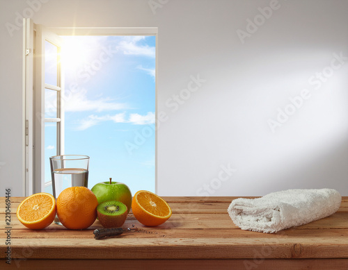 healthy morning wellness concept featuring fresh oranges, green apple, kiwi, a glass of water, and a white towel on a wooden table beside an open window with bright sunshine and blue sky
