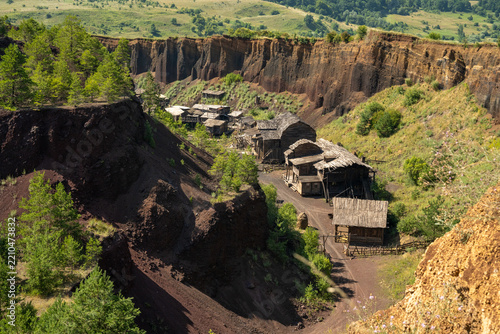 View of rustic wooden structures nestled within the steep, dark red volcanic walls of the Racos scoria quarry, surrounded by pine trees and grassy slopes in Brasov, Brasov, Romania.