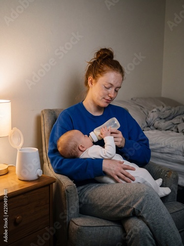 Young mother bottle feeding baby in armchair at night