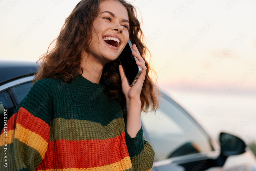 Obraz premium A happy young woman laughs while talking on her phone near a car. Concept for joyful communication, mobile technology, and outdoor lifestyle.