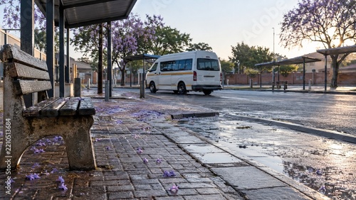 A weathered concrete park bench sits on wet patterned paving stones littered with purple petals near a curb; a white passenger van is parked on the wet asphalt street under an overcast sky