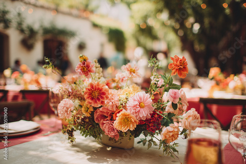 Vibrant floral arrangement featuring pink, orange, and yellow flowers is displayed table wedding. setting is lively and festive, with soft lighting and greenery background, creating joyful