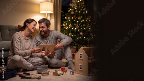 Parents quietly assembling wooden dollhouse late at night with blurred Christmas tree background banner.