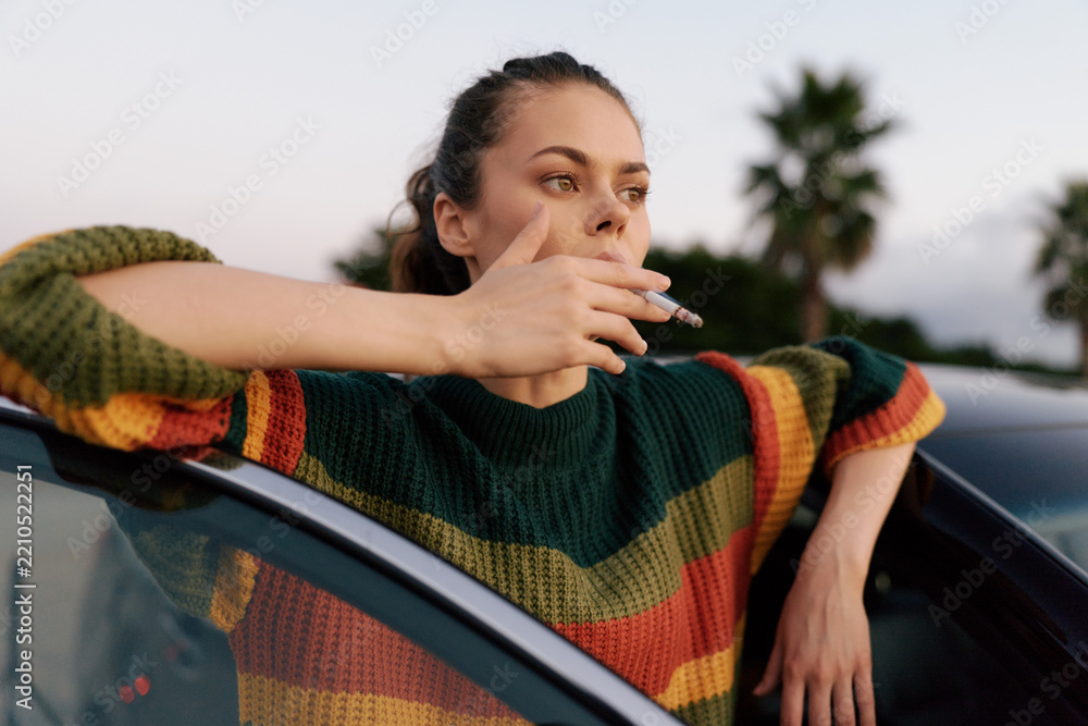 Obraz premium Young woman smoking cigarette leaning on car door outdoors. Casual style and thoughtful mood. Concept of bad habits, lifestyle, and urban youth culture. Soft natural lighting creates a calm