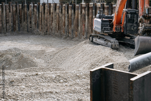 Excavator digging a foundation pit at an urban construction site, heavy machinery working behind temporary sheet piling and safety barriers