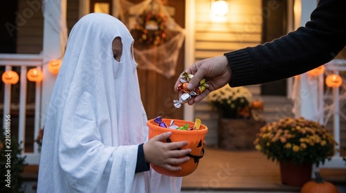 Child in ghost costume trick-or-treating, collecting colorful candy from an adult on a festive Halloween decorated porch