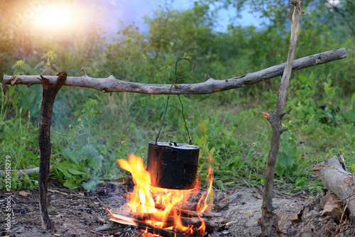 A rustic metal pot hanging over a crackling campfire in a lush green forest wilderness setting