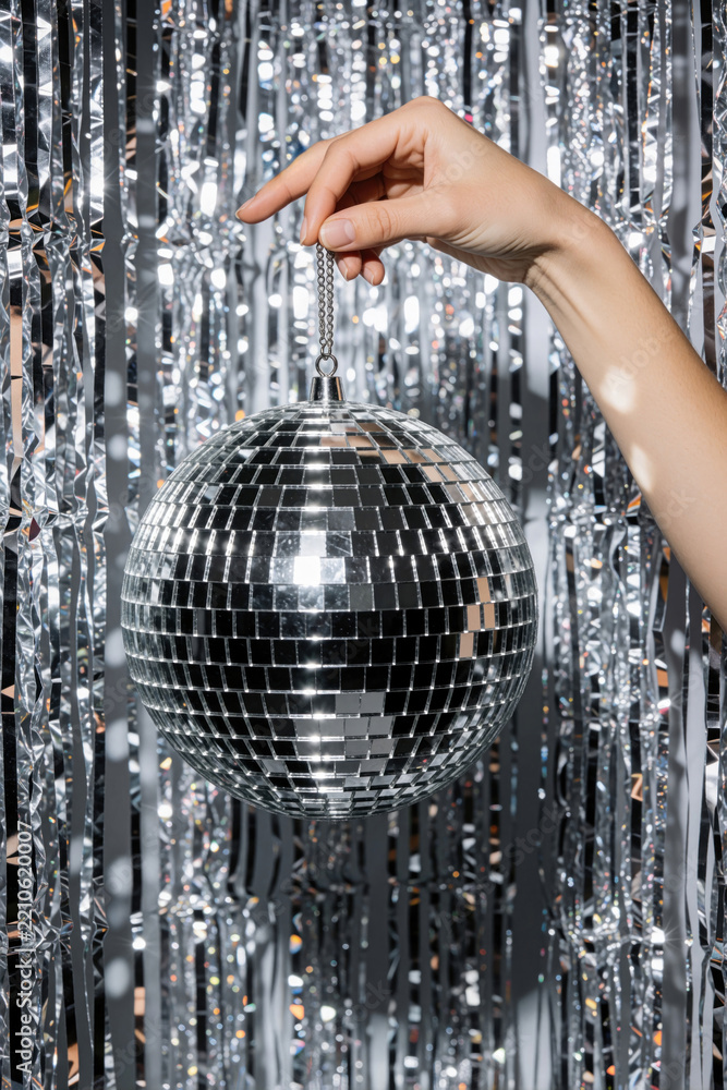 Obraz premium Female hand holds shiny silver disco ball against glittery tinsel background. Woman holds mirrored sphere for party decoration. Festive glitter backdrop creates bright light effects. Glamorous
