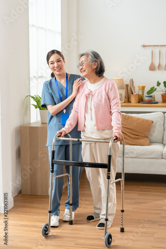 Young Asian nurse assists senior Asian woman practicing walker mobility during rehabilitation, supporting balance, safe movement and growing independence inside modern assisted living center.
