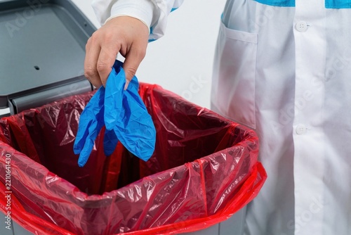 Medical staff discard used blue nitrile gloves into red biohazard waste bin. Doctor in white lab coat performs disposal of infectious medical equipment. Proper hygiene, safety protocols for managing