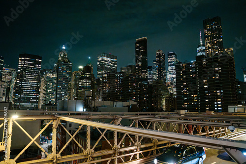 Illuminated skyscrapers of Lower Manhattan rise behind the steel structure of the Brooklyn Bridge at night. Bright city lights and modern towers create a dramatic New York City skyline.