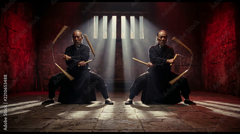 Obraz premium Two martial artists performing synchronized movements with flexible weapons in a stone dungeon, dramatic lighting through window bars. Martial arts training, combat sports, action performance.