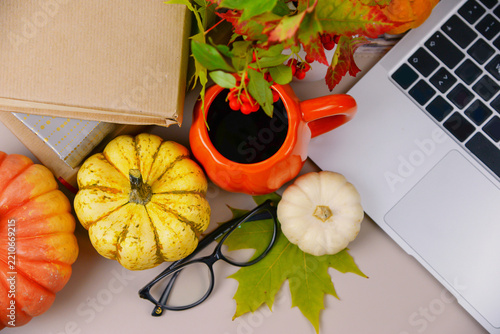 Cozy autumn workspace with laptop, pumpkins, coffee, glasses, books and colorful fall leaves arranged on a neutral desk.