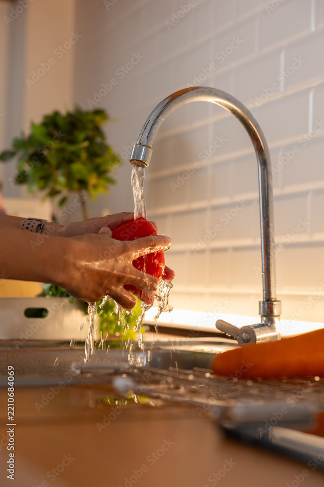 Obraz premium Hands washing fresh red bell pepper under running tap water in modern kitchen sink. Close-up shot showing water splash, cleaning organic vegetables for healthy food cooking preparation Vertical
