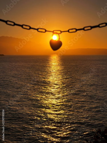 Romantic heart-shaped padlock hanging from a metal chain with the setting sun shining through it over a calm sea, symbolizing love, commitment, romance, and lasting relationships.