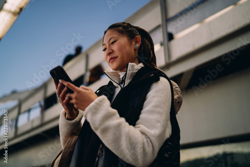 Asian woman looking at smartphone screen, digital nomad lifestyle, mobile application, technology connection, authentic representation, inclusion, real people, urban setting, communication, online
