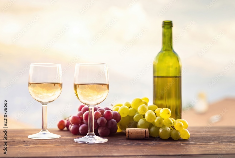 Obraz premium Wine glasses and bottle on wooden table. Fresh red and green grapes with cork. Sunset vineyard background